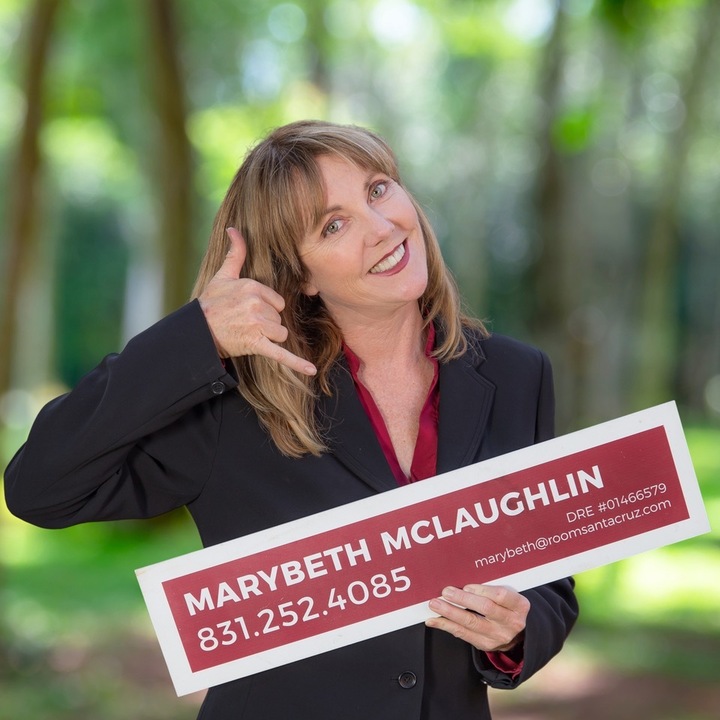 A headshot of MaryBeth using a fingers as a call a phone by her ear and mouth to call her. She is holding her sign with her info on it.